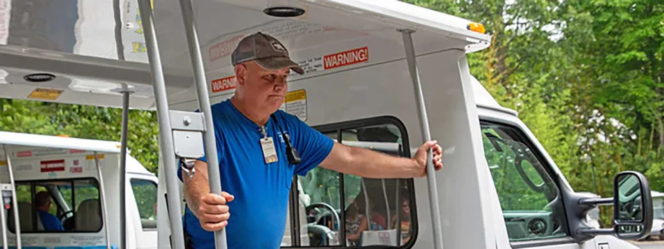 An older, light skinned man wearing a brown hat, blue t-shirt, and khaki shorts, stands in the back of a white tram, between two rows of chairs and resting his hands on two poles that stand on either side of him. Trees are visible in the background and there is a North Carolina Zoo logo on the side of the white tram door.