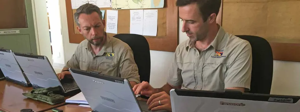 Two men, both wearing light brown short-sleeved shirts with a NC Zoo logo, are sitting side-by-side at a table working on laptop computers. The men are focused on their screens, typing. In the background, there is a corkboard wall with maps and documents pinned up.