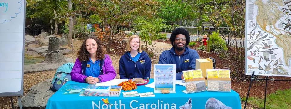 Three Zoo educators smile while seated behind a table at the Zoo's scenic outdoor playarea known as Kidzone. The table is draped in a bright blue tablecloth featuring the Zoo's logo and assorted wildlife. The table contains educational materials, drawings, seeds, and maps featuring birds.