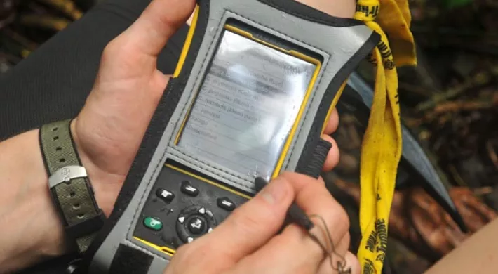 View of someone's hands holding a SMART spacial monitoring and reporting tool, which is a small, black handheld device the size of a cell phone.
