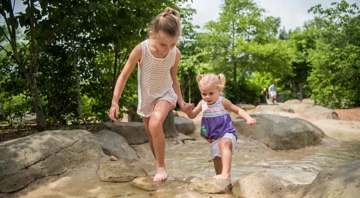 A young girl holding hands with a toddler as they climb barefoot up a steep, rocky hill. They are surrounded by trees and large rocks.