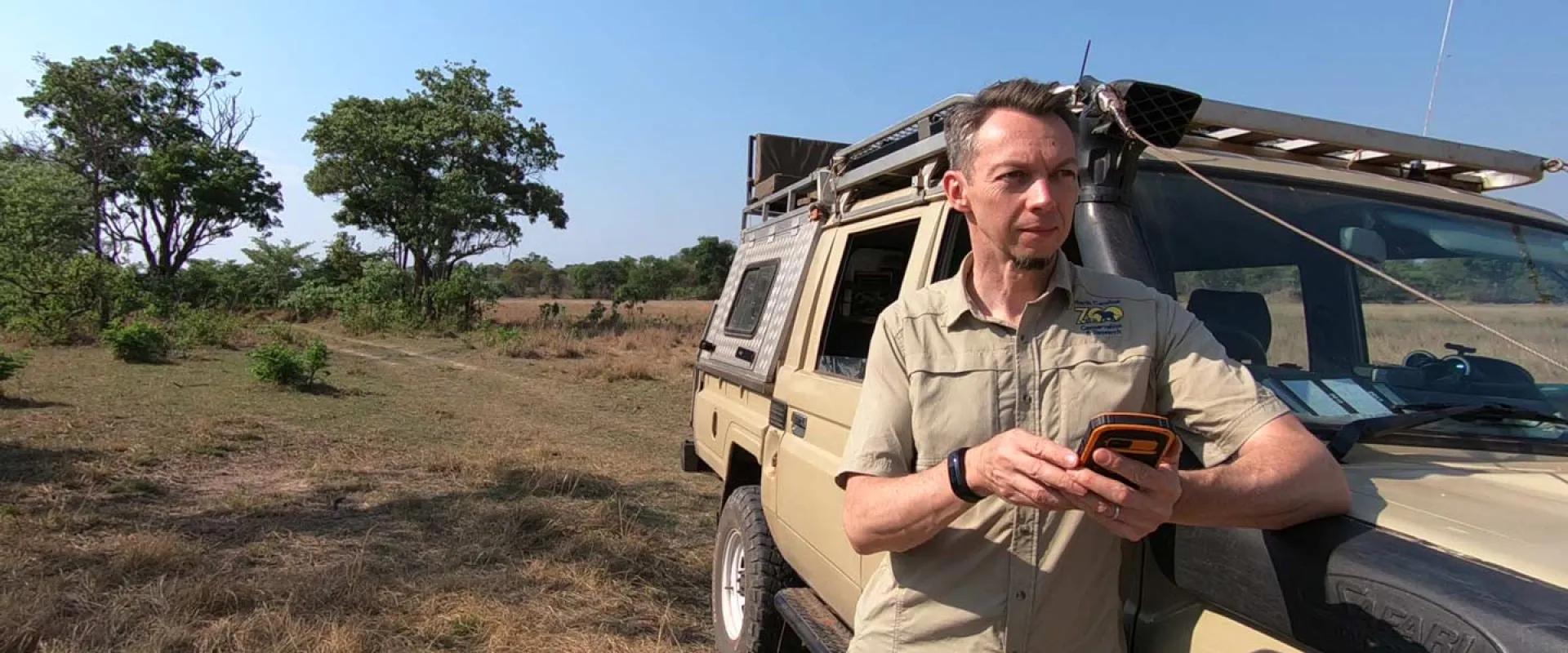 A man in a light brown shirt stands by a tan off-road vehicle in a savanna-like setting, looking at a dark-colored phone. The background shows sparse vegetation, green trees, and a clear blue sky.