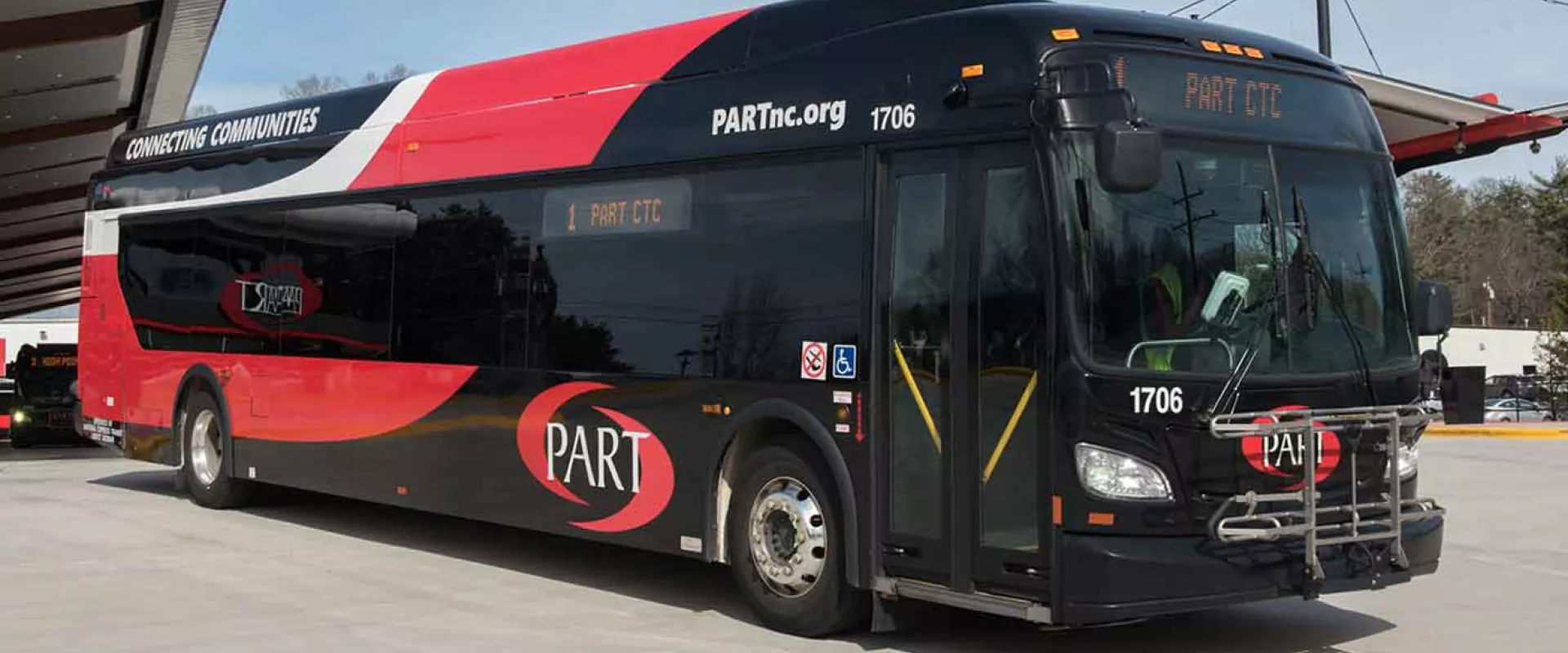 A large bus with a red and black design on the side with the word "PART" on it parked in front of a bus shelter with a slanted metal roof. There are flag poles off to the left with half mast flags.