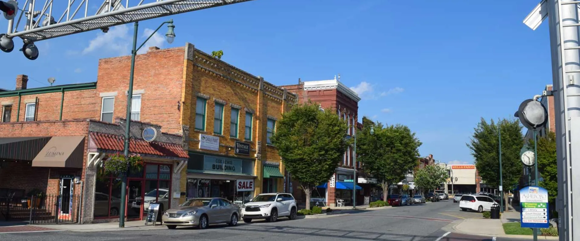 A small town square with a narrow street that is lined with quaint brick buildings and cars parked along the sides. Trees are spaced evenly down the sidewalks under a bright blue sky.
