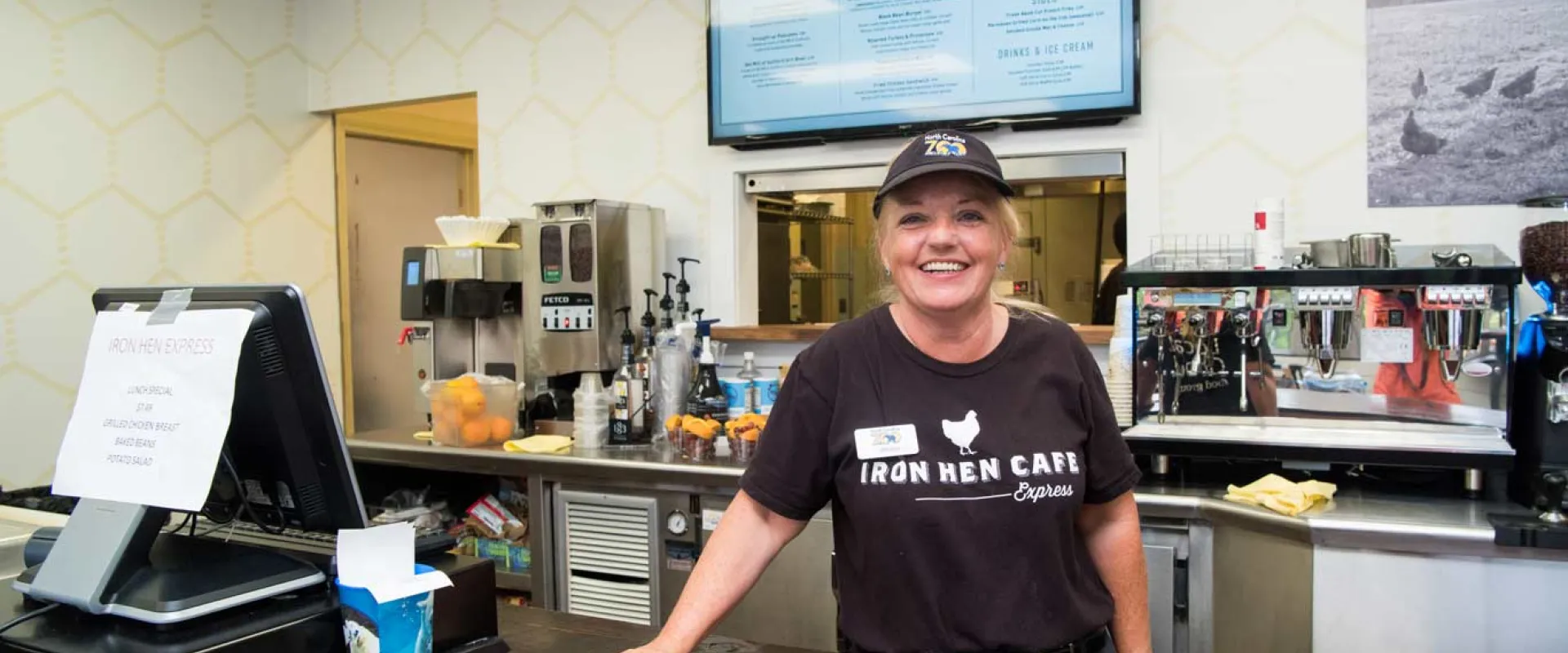A woman wearing an Iron Hen Cafe hat and uniform leans casually on a wooden restaurant counter next to a cash register. There are food items, kitchen machinery, and kitchen visible behind her. A menu hangs on the wall behind her head.
