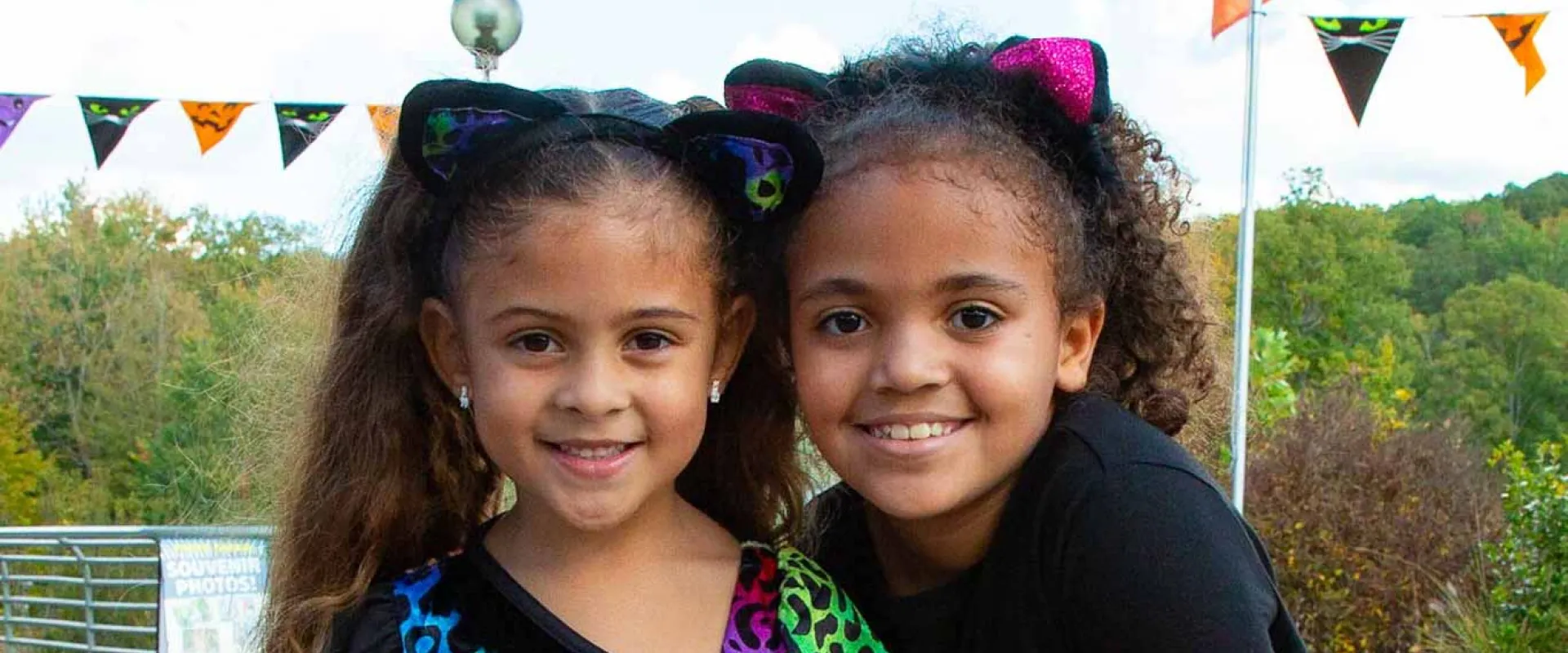A close-up of two young girls smiling and standing close together. Both are wearing black cat ear headbands with cat ears. The girl on the left has long, straight hair and is wearing a black shirt with a bright, multi-colored animal print. The girl on the right has curly hair and is wearing a solid black shirt. In the background, there is a string of orange and black Halloween flags, and a landscape of trees under a cloudy sky.