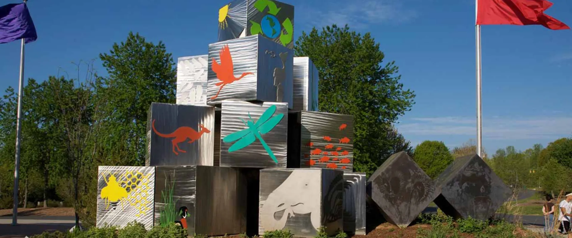 A striking silver cube sculpture, representing themes of conservation and nature, stands on a grassy mound near a pathway. The cubes are adorned with brightly colored environmental and wildlife graphics.