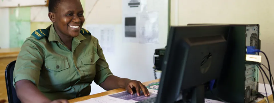 A person appearing to be a woman with short dark hair, possibly a ranger given the green uniform with yellow and black shoulder markings, is seated at a desk. She is smiling and looking towards a computer monitor. Her hands are positioned on a computer mouse and keyboard. Behind her, there is a map on the wall and some papers. A computer tower and monitor are visible on the desk.