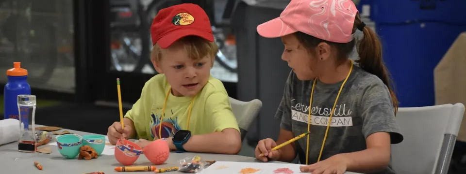 Two children sitting at a fold out table with paper, pencils, crayons, plastic eggs, and water bottles, doing a camp craft. The child on the left is a boy with short blonde hair and a red baseball hat, and to the left is a girl with a dark pony tail and a pink baseball hat.