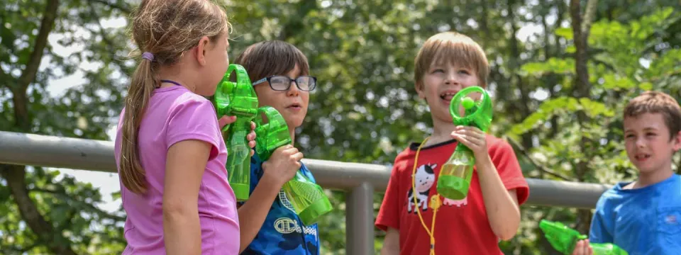 A group of four young campers keeping cool outside with green misting fans. There is a metal handrail behind them and trees in the background.