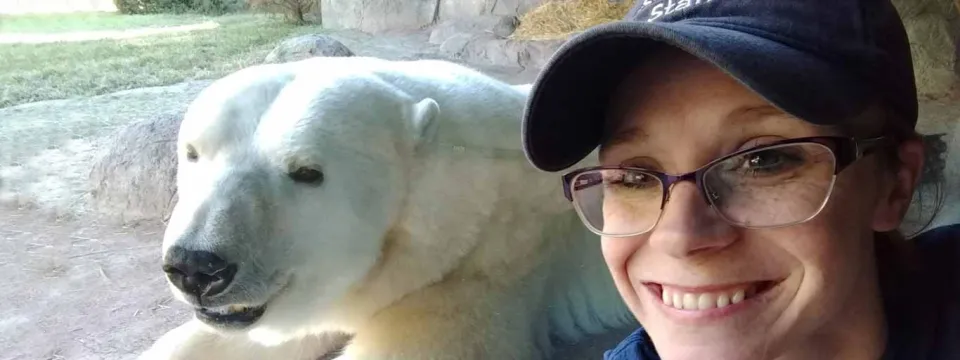 A smiling woman with glasses and a baseball cap is taking a selfie with a polar bear resting behind her. The woman is on the right side of the photo, and the polar bear is on the left.