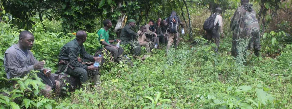 A team of approximately eight men, many wearing green and camouflage uniforms, takes a break and sits among dense green undergrowth in a rainforest or jungle.