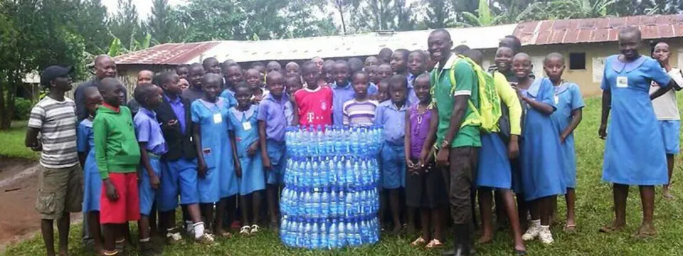 A group of approximately 30 students in light blue uniforms, along with a few adults, are posing with a tall stack of blue bucket-style water filters outdoors on a grassy field.
