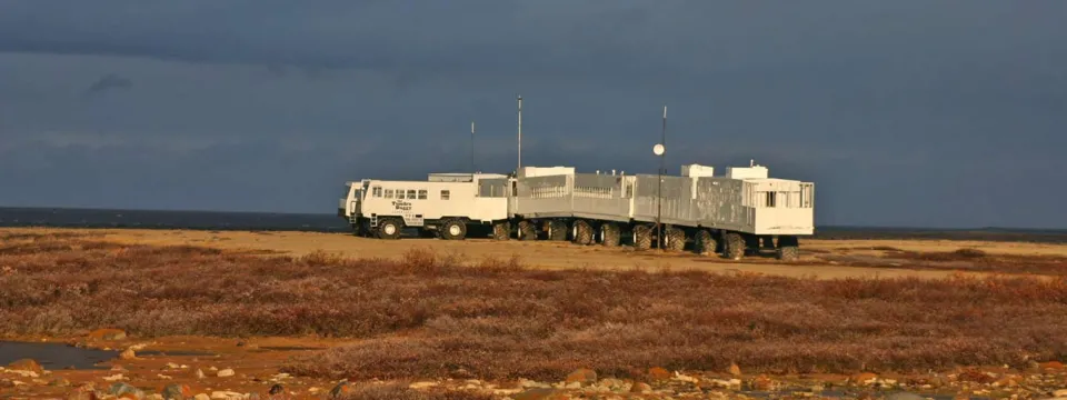 A long, white bus or transport vehicle driving across a vast, flat expanse of reddish-brown desert or outback ground. The ground takes up the majority of the frame and appears barren. A dark, overcast or stormy sky dominates the background, creating a sharp contrast with the landscape and the vehicle.