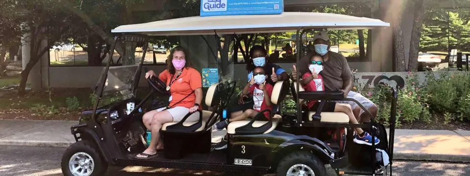 Several people, including a man in an orange shirt driving, are seated in a black, four-seater golf cart with a white roof and a blue sign attached to the top, driving through a shaded, outdoor area.