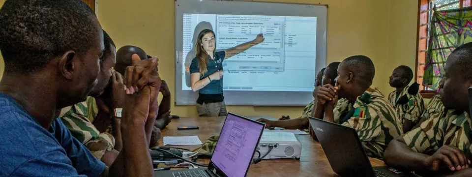 An indoor training session with a woman in a polo shirt pointing to a data spreadsheet projected onto a screen. She is instructing a group of about eight men seated around a table, all wearing camouflage uniforms. Several laptops are open on the table.