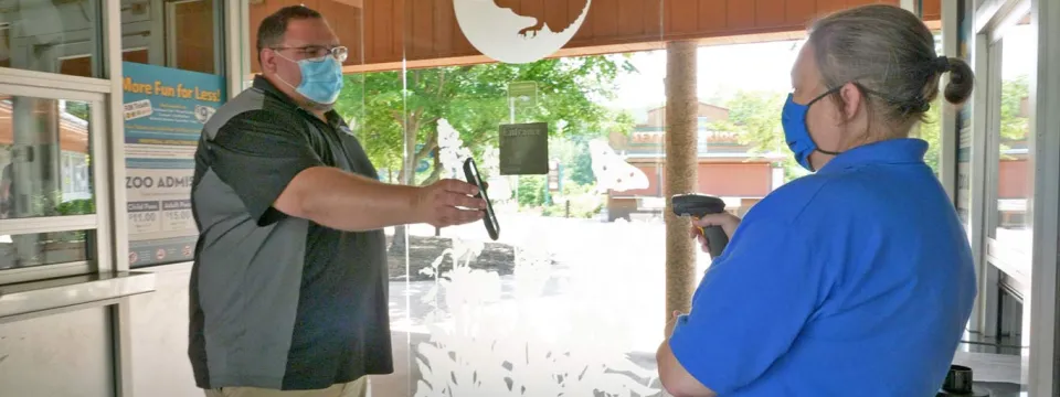 Two adults wearing face masks and standing a safe distance apart near a doorway. The man on the left, wearing a grey and black collared shirt and tan pants, holds his arms out in front of him showing something. The person on the right, wearing a bright blue shirt and a blue face mask, is facing him. The background includes an open doorway leading outside to visible trees and a white, stylized logo of a circular shape on the wall. The scene suggests a modern retail setting during the COVID-19 pandemic.