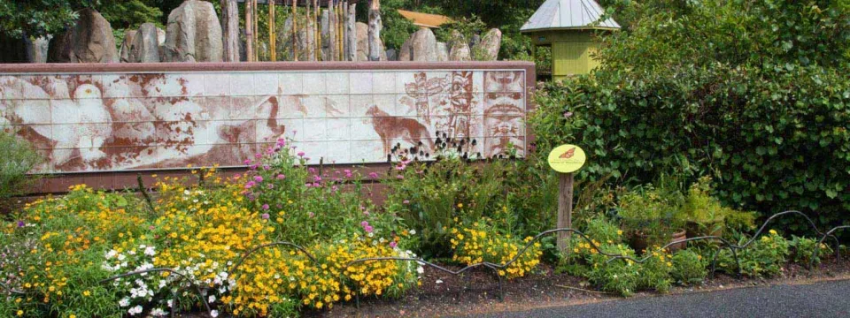 An outdoor zoo exhibit sign is set amidst lush landscaping and trees. The main sign is a long, rectangular structure with a painted panel featuring images of animals, including a small cat-like animal visible on the right. The sign is surrounded in the foreground by colorful flower beds with yellow, white, and pink blooms. In the background, there are large vertical stone pillars and a small structure with a conical metal roof.