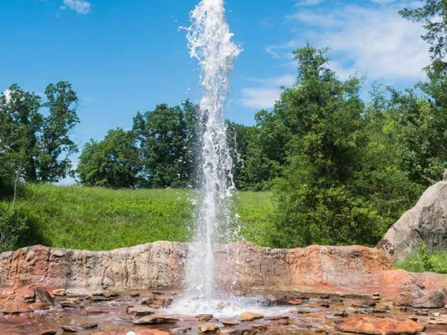 A fountain or geyser feature spraying a tall column of water high into the air against a bright blue sky with scattered white clouds. The water erupts from a base of reddish-brown and tan rocks, which are wet and surrounded by smaller, dark stones. The background is a vibrant green landscape with lush trees and a grassy hillside under the sunny sky.