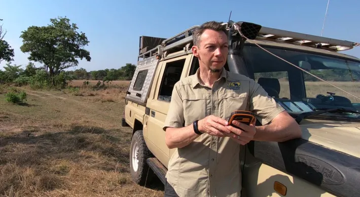A man in a light brown shirt stands by a tan off-road vehicle in a savanna-like setting, looking at a dark-colored phone. The background shows sparse vegetation, green trees, and a clear blue sky.