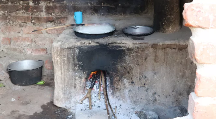 A traditional earthen cookstove, is shown with two pots on top. Flames are visible inside the firebox. A blue mug and a metal bucket are placed nearby. The cookstove is constructed from mud and bricks and appears to be inside a brick room.