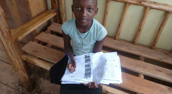 A young child sits on a wooden bench, holding a notebook and pen and looking up at the camera. They have dark skin, short hair, and are wearing a light blue shirt and a black skirt.