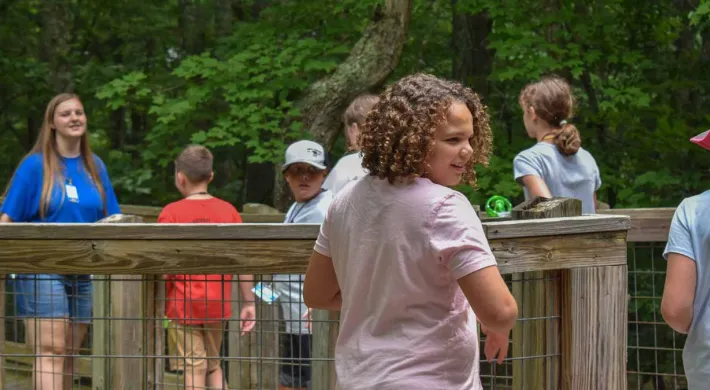 A group of six children walking in a line behind a woman with long hair and a blue uniform shirt, over a wooden bridge through a forest. They are facing her, and she is turned and walking backwards, seemingly to talk to them.