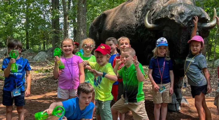 A group of young campers holding green personal fans stand together, posing in front of a large brass statue of a Bison in a wooded area.
