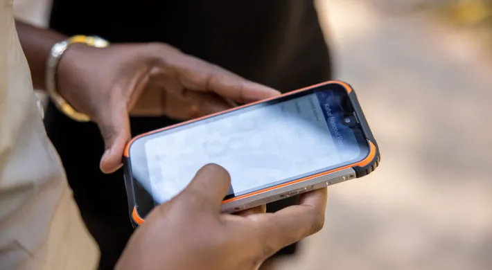 A pair of hands holding a device that looks like a cellphone. Another person, visible from the waist down, wearing black pants, is standing next to them.