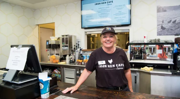 A woman wearing an Iron Hen Cafe hat and uniform leans casually on a wooden restaurant counter next to a cash register. There are food items, kitchen machinery, and kitchen visible behind her. A menu hangs on the wall behind her head.