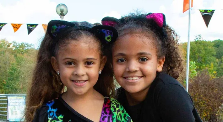 A close-up of two young girls smiling and standing close together. Both are wearing black cat ear headbands with cat ears. The girl on the left has long, straight hair and is wearing a black shirt with a bright, multi-colored animal print. The girl on the right has curly hair and is wearing a solid black shirt. In the background, there is a string of orange and black Halloween flags, and a landscape of trees under a cloudy sky.