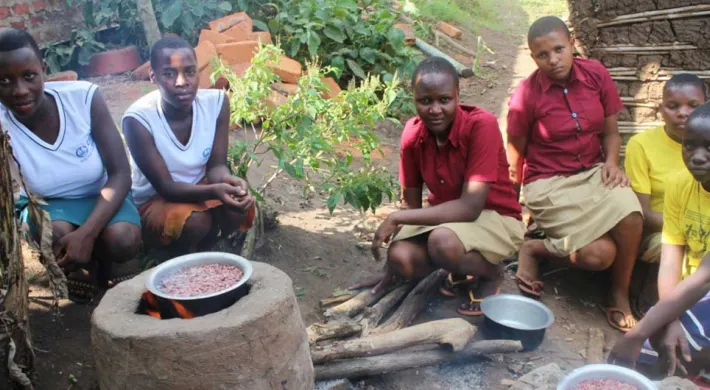 Six young women or girls are crouching around a traditional outdoor cooking fire set up on the ground, likely in a rural African community. In the center, a cooking pot filled with red beans sits on a raised clay or stone stove. The girls are looking towards the viewer, wearing a mix of school uniforms (white vests and skirts/wraps, or maroon shirts) and yellow t-shirts. More cooking pots and wood are visible on the ground.