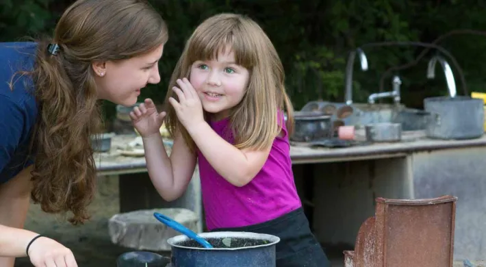 An Educator is talking with a young white girl in pink at the Mud Cafe with a steel counter behind her that is littered with kitchen utensils.