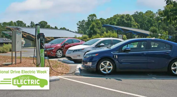 Three electric cars, a blue, white, and red sedan, are parked at a charging station with solar panels visible in the background. A large green and white logo for "National Drive Electric Week" is in the lower left corner.