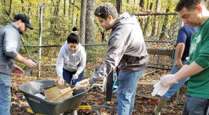A group of five volunteers is working outdoors in a wooded area surrounded by a chain-link fence, loading various items, including cardboard, into a wheelbarrow.