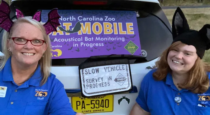 Two smiling women wearing blue shirts and novelty animal ear headbands (one with purple bat wings, one with black pointed ears) stand in front of the open trunk of a vehicle. A large purple and yellow sign reading "MOBILE" is visible in the trunk, likely for an outreach or education program.