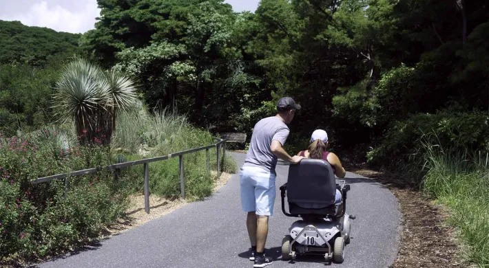 A man wearing a grey shirt and light-colored shorts is standing on a paved pathway, gently guiding or assisting a person seated in a powered wheelchair. The path is surrounded by lush green vegetation, including a unique Yacca plant on the left side, behind a low metal railing.