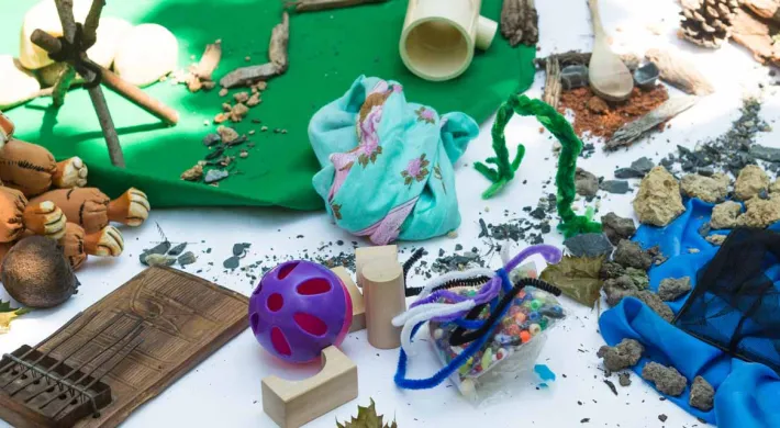 A table covered in a white cloth and littered with a variety of craft items including a toy tiger, green clay, fuzzy pipecleaners, pinecones, green paper, sand, sticks, wooden blocks, and a blue plastic material.