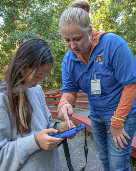 Educator with a young Asian girl showing her how to use a program on a tablet. There are wooden benches and trees behind them, giving the impression of an outdoor ampitheatre.