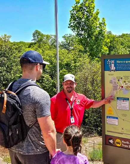 Volunteer standing beside a sign with a large Zoo map talking with a man and his young daugher
