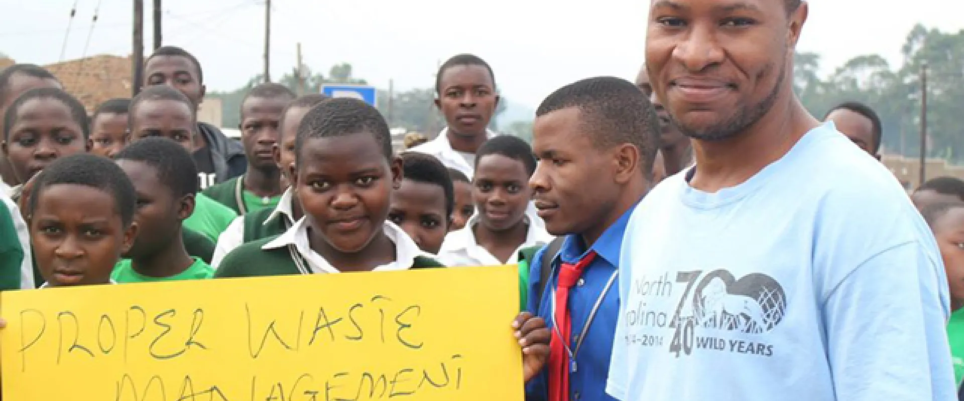 Bruce, a young, dark-skinned man wearing a blue NC Zoo shirt smiles as he stands with a group of dark skinned children of varying ages from with Bigodi secondary school club. Two of the children hold a yellow sign with the words, "Proper waste management is everyone's responsibility" written in black marker.