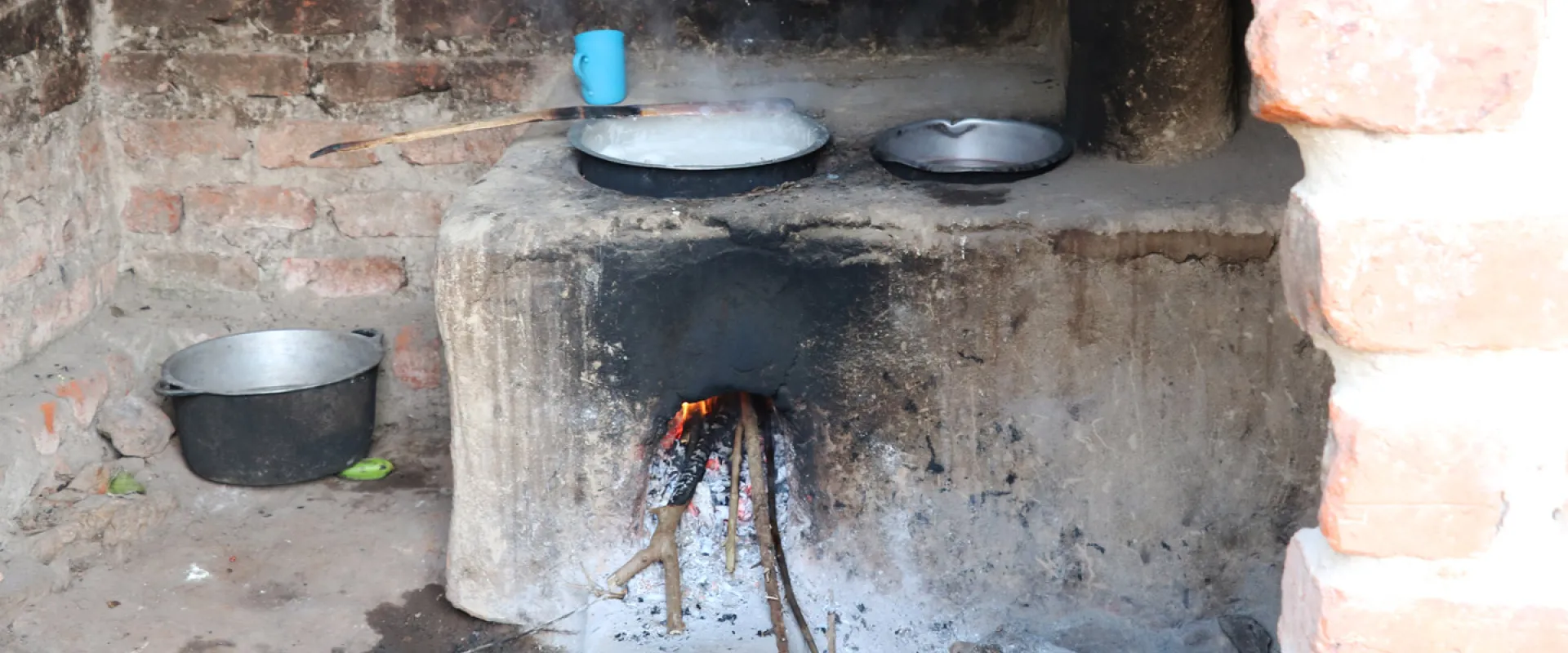 A traditional earthen cookstove, is shown with two pots on top. Flames are visible inside the firebox. A blue mug and a metal bucket are placed nearby. The cookstove is constructed from mud and bricks and appears to be inside a brick room.