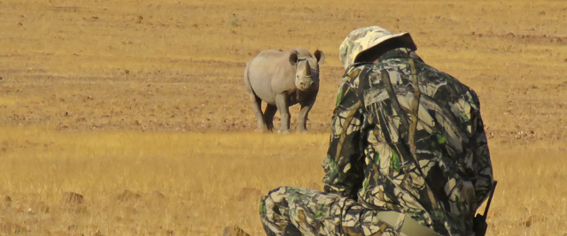A person wearing camouflaged colored clothes crouched down conducting a rhino monitoring with Save the Rhino Trust. There is a rhinoceros in the background facing the person in a savanna environment.
