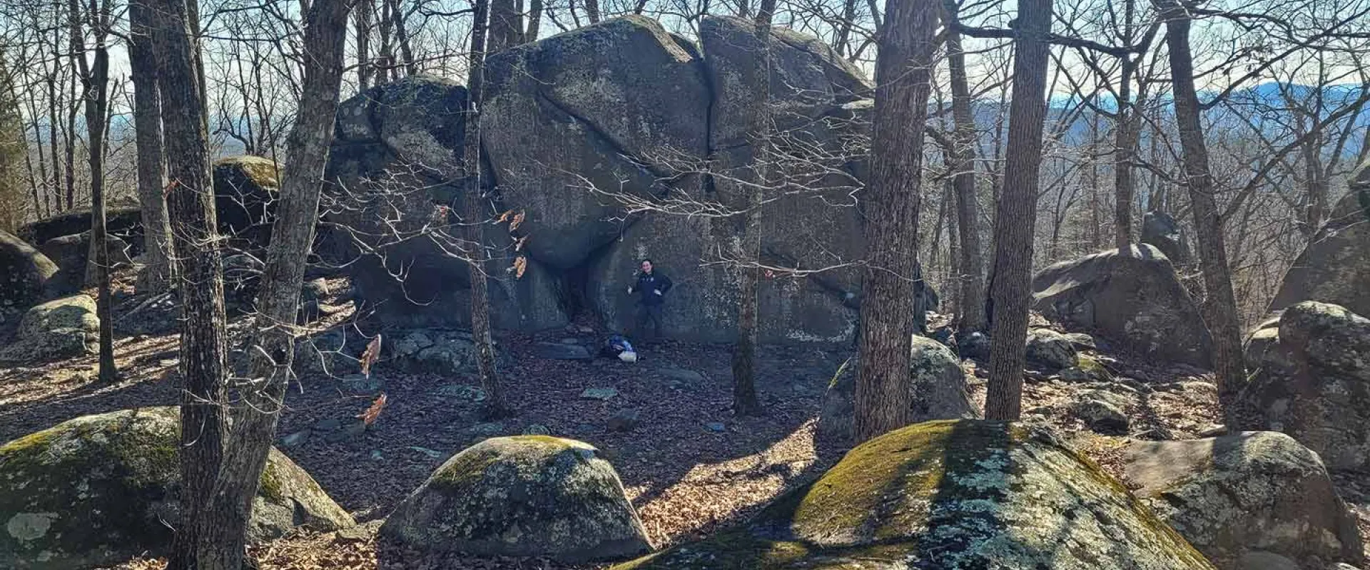 A wide shot shows a wooded area, likely Ridges Mountain Nature Preserve, featuring large, moss-covered rocks and bare trees. A person is visible among the rocks.