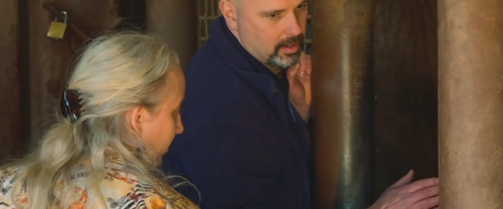 Two adults in a North Carolina Zoo enclosure examine an Elephants leg. One, with long blonde hair and an animal-print shirt, holds up a small pin. The other, bald with a beard and in a blue shirt, looks left and touches the leg gently.