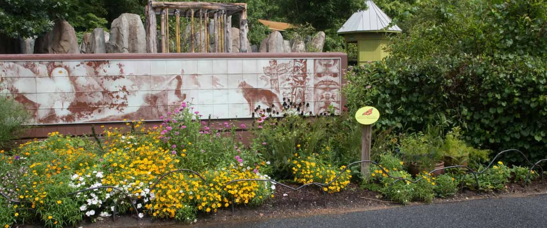 A low-angle shot captures a section of a zoo exhibit. In the foreground, a bed of yellow and white flowers borders a dark asphalt path. Behind them is a long, horizontal mural depicts what appears to be various animals in a rustic, almost weathered style. Above and behind the sign, a structure resembling ancient ruins with multiple stone columns is visible. The background is filled with a thick canopy of green trees.