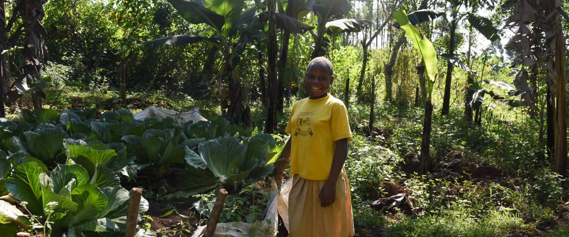 A person with dark skin and a shaved head stands smiling in a sunny, lush green garden. They are wearing a bright yellow t-shirt and a tan skirt. To their left, large, leafy green plants are growing in rows in some light-colored sacks that have been repurposed for gardening. Tall trees and various green plants fill the background, suggesting a tropical environment.