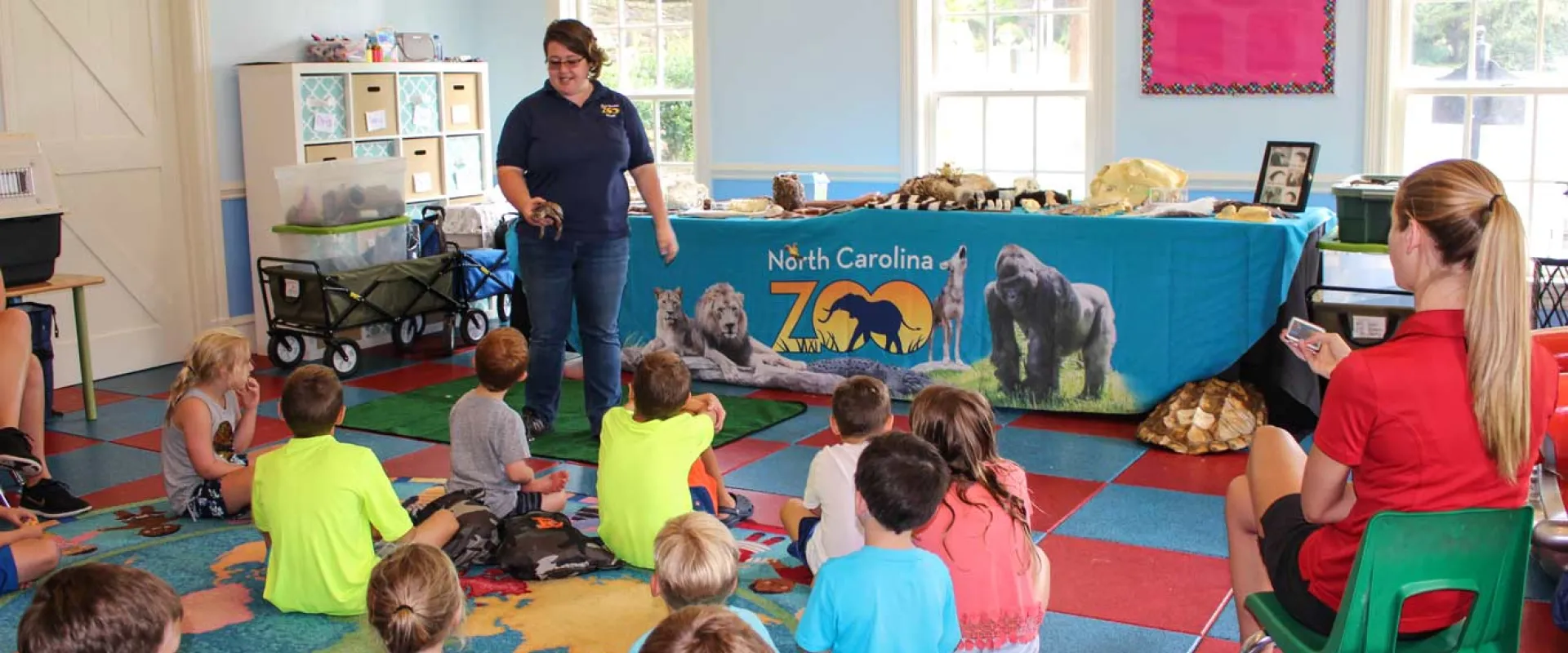 A woman in a navy NC Zoo uniform stands with a long table, covered with a teal NC Zoo tablecloth and various animal artifacts. In front of her is a group of children seated on colorful floor tiles, and a woman with a long blonde ponytail and red collared shirt sitting in a green chair to the right. The setting appears to be an elementary school classroom.