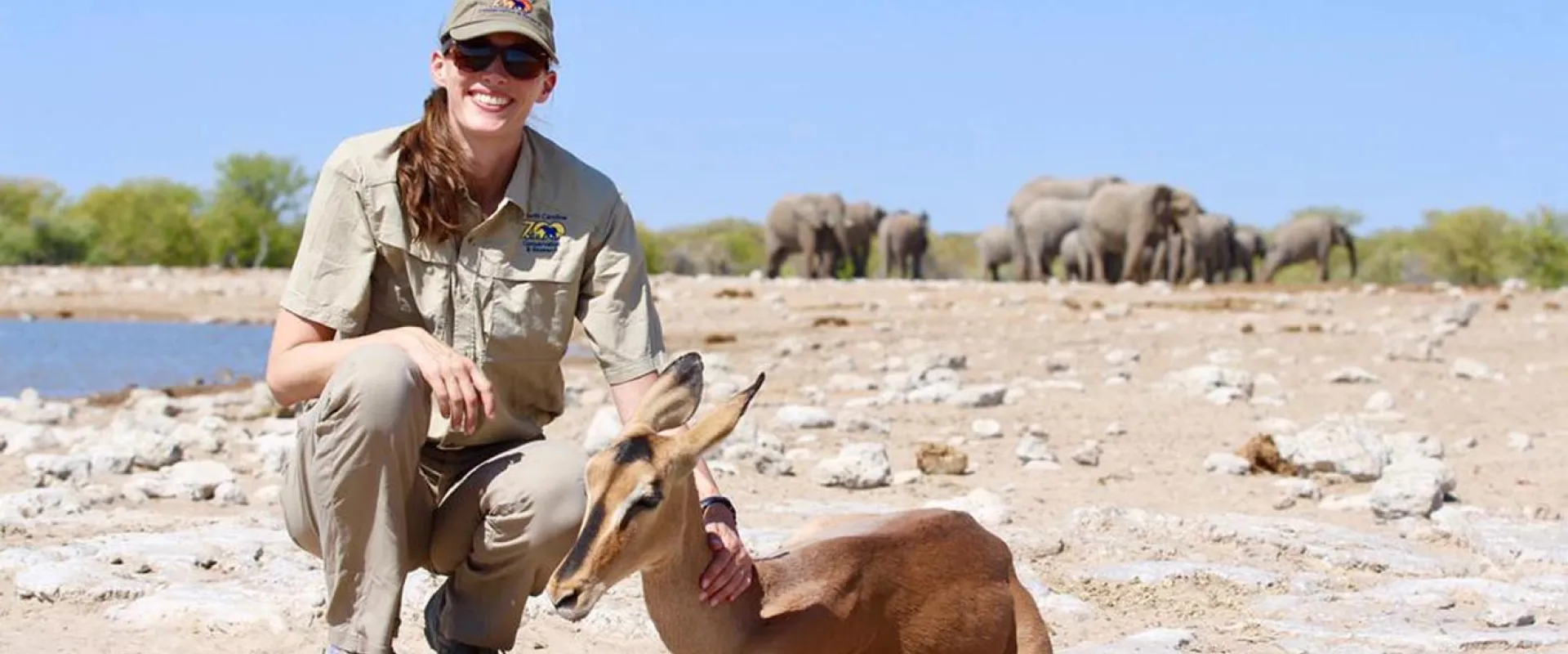 A woman in a tan rangers uniform squats smiling with a hand resting on the long delicate neck of an Addra Gazelle lying next to her. The two are on dry, rocky soil next to a small lake. A herd of Elephants can be seen in the far distance.