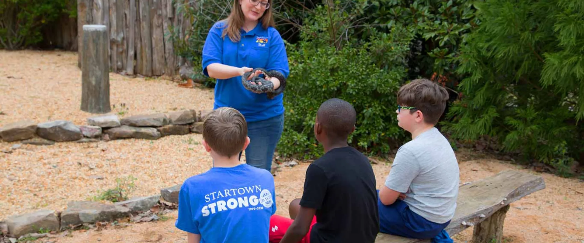 A nature educator, a woman with glasses and a blue uniform shirt, is holding a dark snake while presenting to three young boys seated on a wooden bench. The setting appears to be an outdoor educational area, possibly at a zoo or nature center, with a dirt ground and a wooden fence/structure in the background.